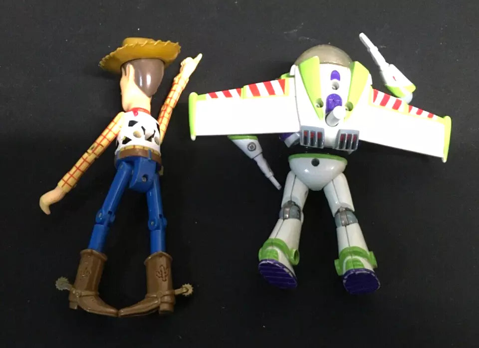 Back view of Toy Story Woody and Buzz Lightyear action figures lying side by side. Woody is wearing his signature cowboy outfit with boots and a hat, while Buzz Lightyear has his wings extended with red and white stripes. Both figures show signs of play wear."