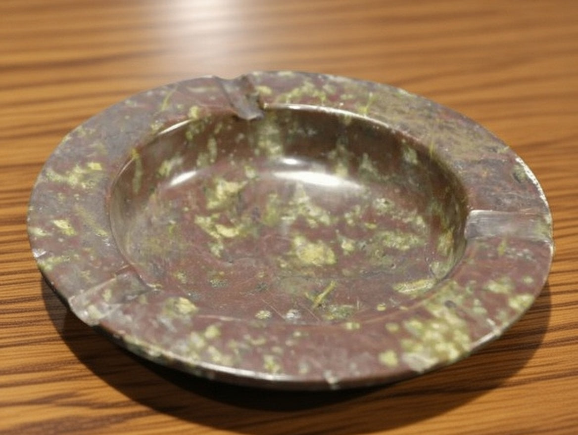 Polished natural stone ashtray on wooden surface, brown rock smoking tray with green speckled pattern and three cigarette rests