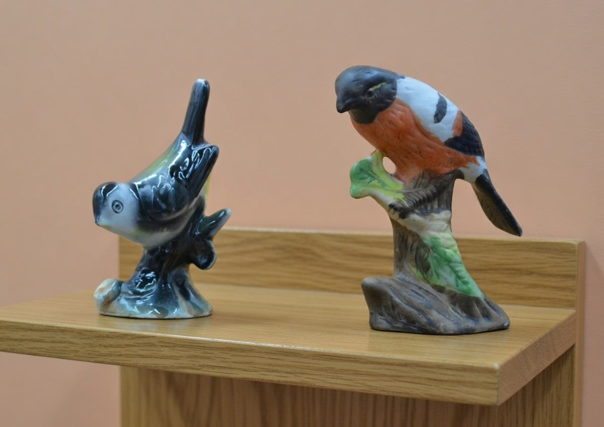 Pair of vintage ceramic bird figurines, one black and white bird in flight and one bullfinch perched on a branch, hand-painted gloss finish, displayed on wooden shelf, collectible ornaments in good condition