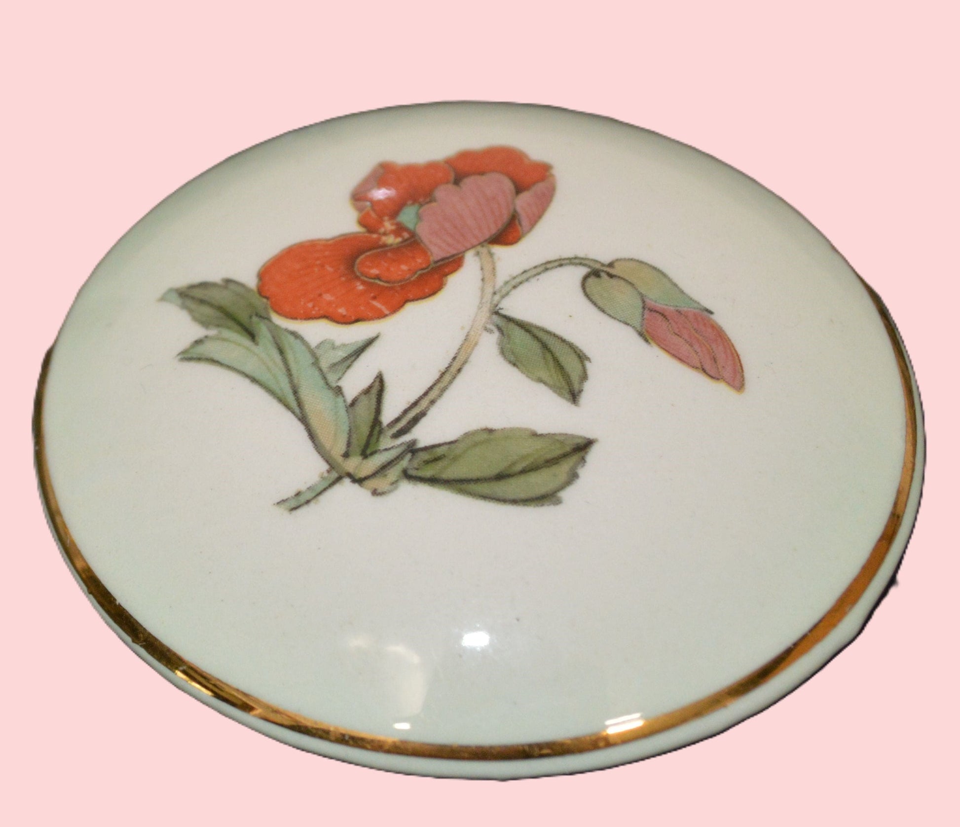 The ceramic lid of the box is shown, with the red flower and green leaves artwork clearly visible. The flower has a distinct orange-red color, and the lid is placed on a green surface.