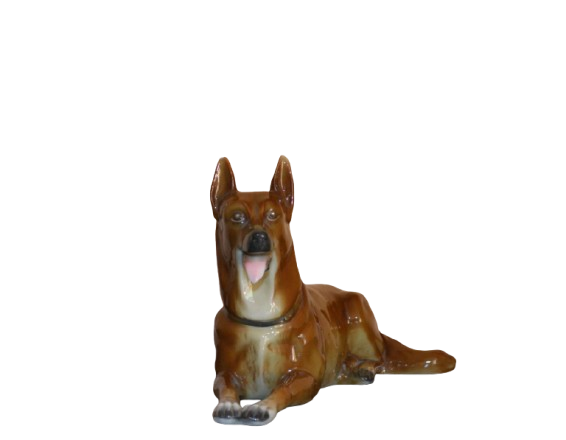 Ceramic figurine of a brown German Shepherd or similar dog, lying down with its front paws extended and mouth slightly open, showing a pink tongue