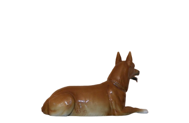 Side view of a ceramic dog figurine with perked ears, an open mouth, and a glossy brown finish. The dog is lying down with its front legs extended and hind legs tucked under."