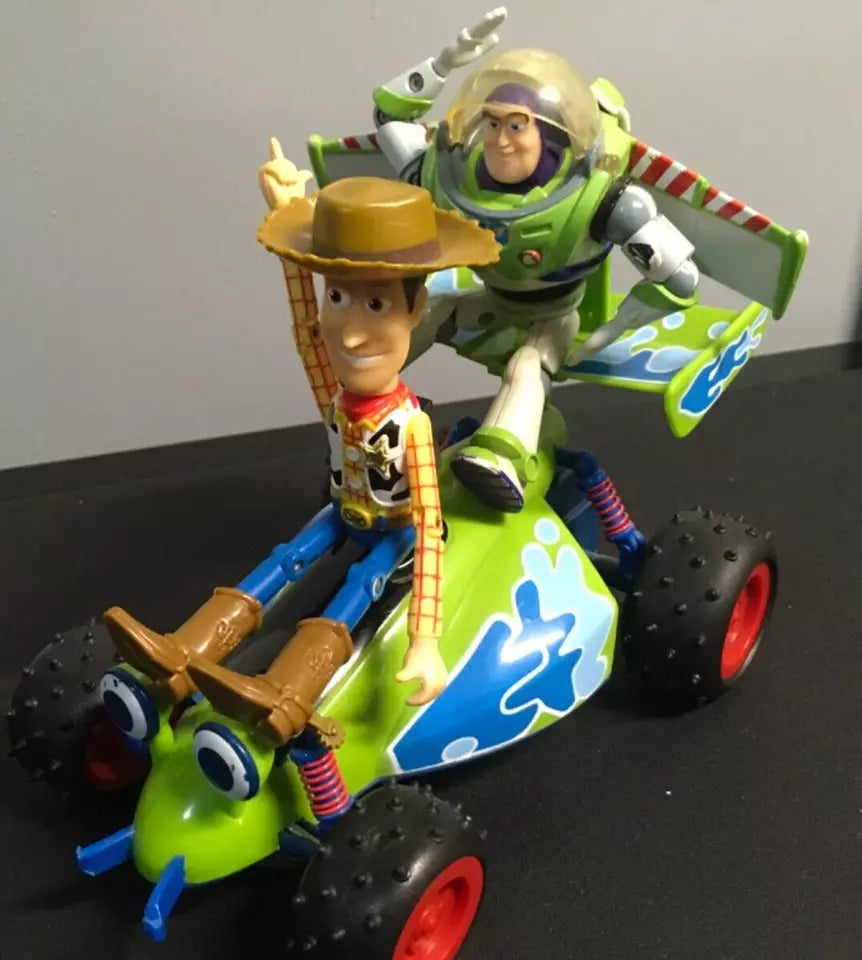 Toy Story Woody and Buzz Lightyear action figures riding a free-wheeling RC car. Woody sits at the front with his arm raised, while Buzz is positioned at the back with his wings extended. The RC car features a vibrant green and blue design with red wheels and expressive eyes on the front. The figures and vehicle show signs of play wear."