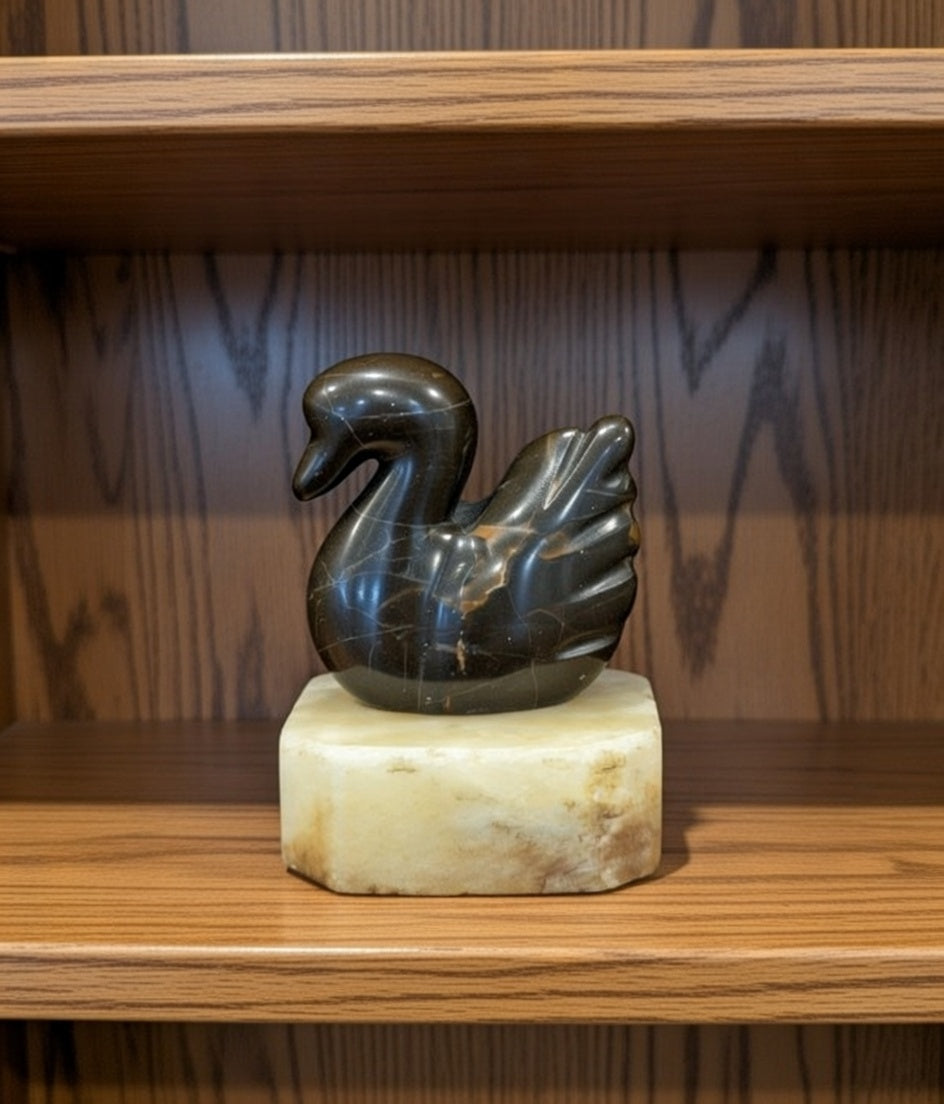 Polished dark stone swan figurine with natural veining displayed on a white marble base, decorative natural rock bird ornament on wooden shelf.