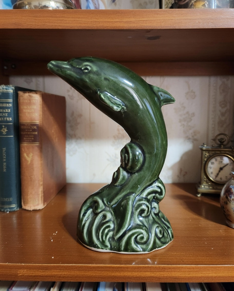 Vintage green ceramic dolphin figurine on wave base displayed on wooden shelf with books and decorative clock